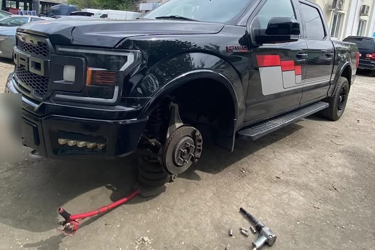 Camionetă ridicată pe cric în curtea atelierului, cu roata din față scoasă: se văd discul de frână și butucul, iar pe jos cheia pneumatică și prezoanele.
